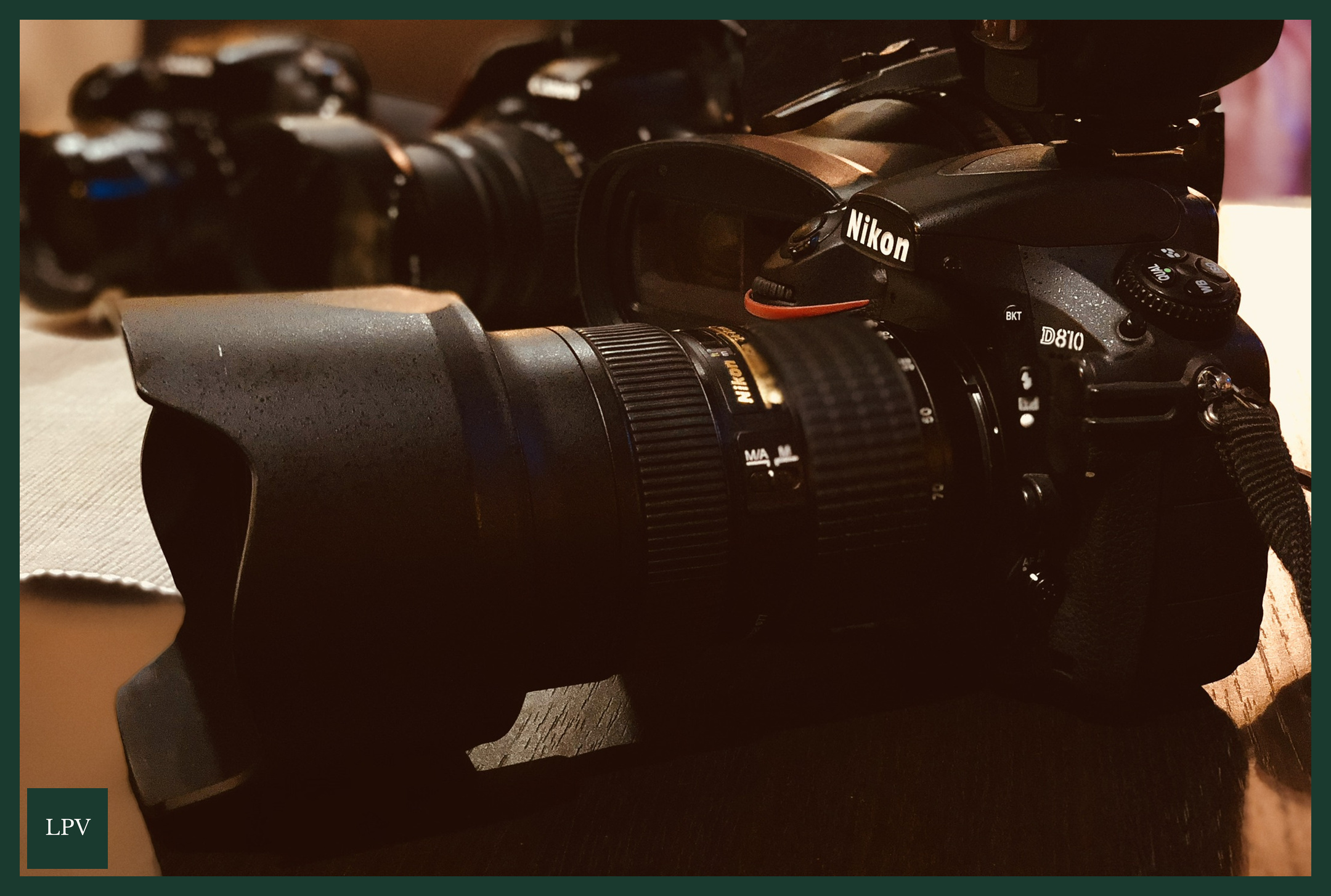 Nikon D810 with long lens and hood under warm light beside a blurred camera.